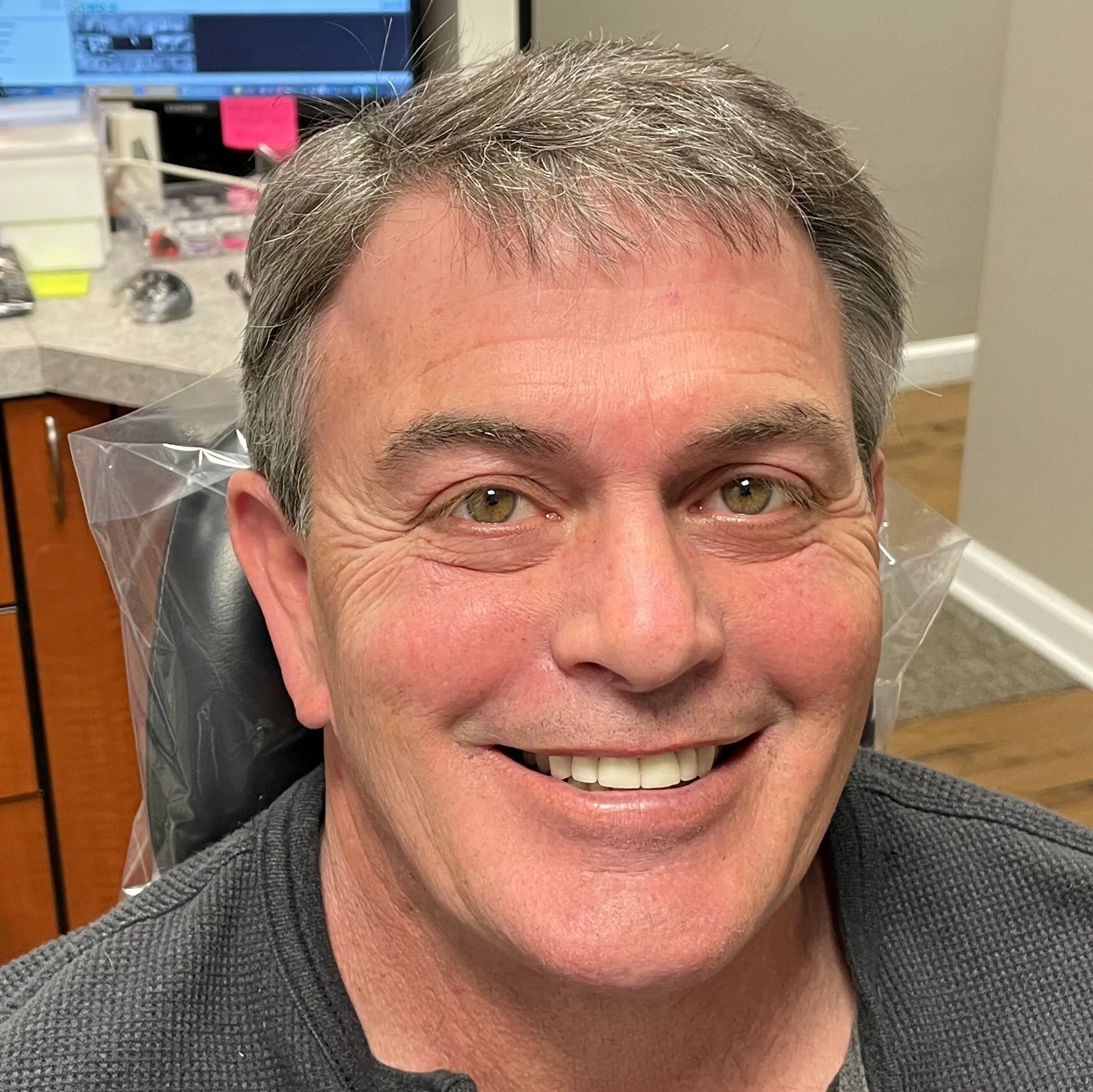 Smiling man in gray shirt sitting in dental office with desk and supplies visible.