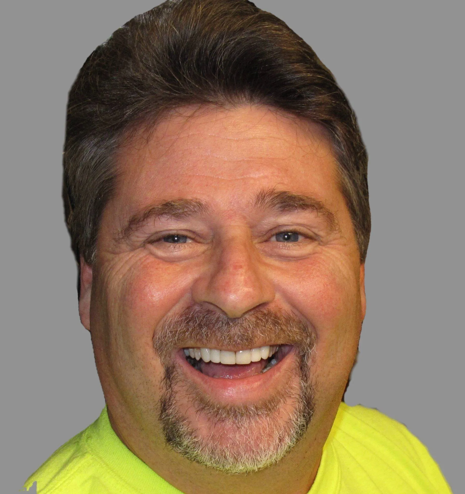 Smiling male dentist in yellow-green scrubs against gray background, professional headshot.