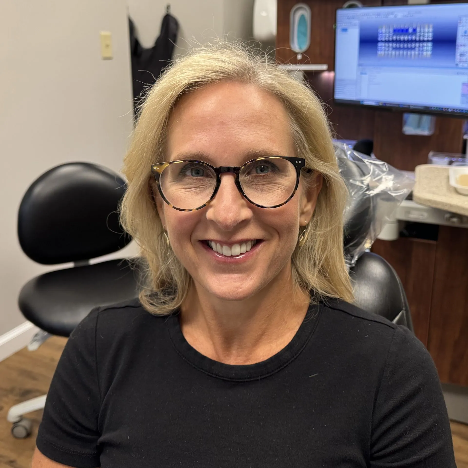 Woman wearing glasses smiling at camera in dental office with computer monitor visible behind her.