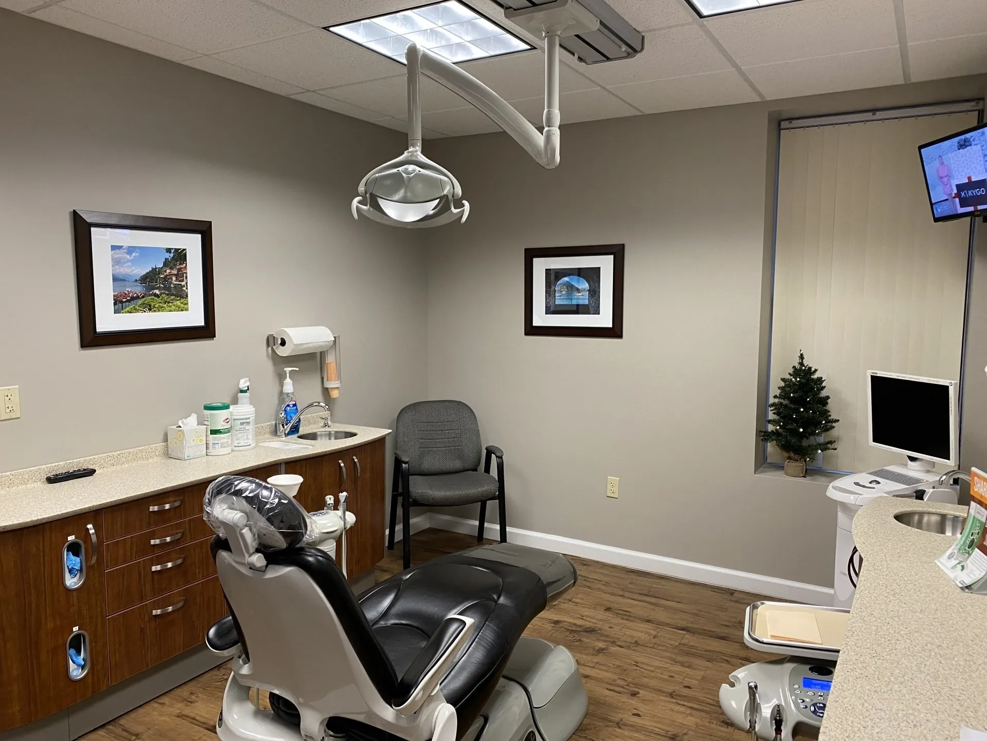 Modern dental operatory with patient chair, overhead lighting, wooden cabinetry, and framed artwork on walls.