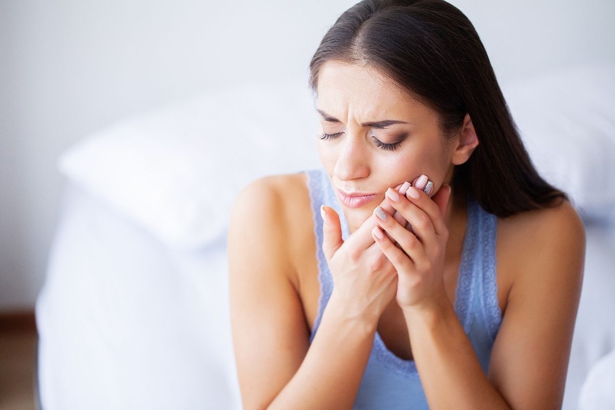 Woman holding her cheek in pain, appearing to experience tooth or jaw discomfort indoors.